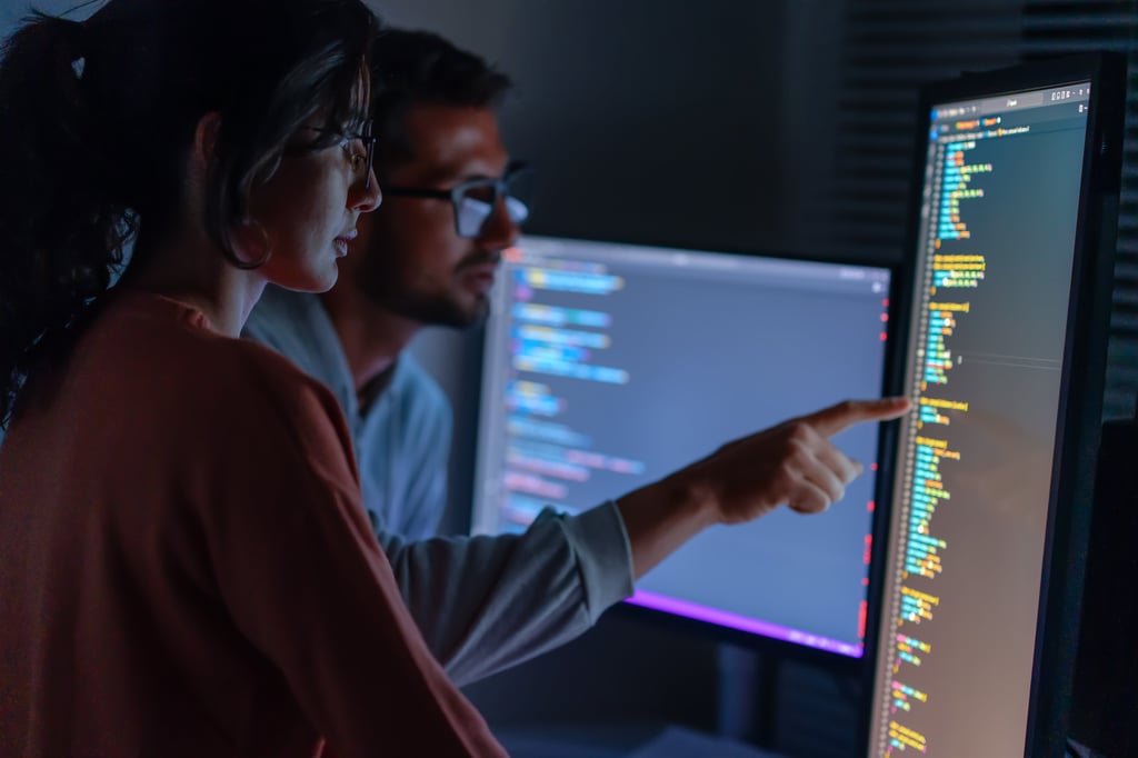 Two software developers collaborating on a coding project, analyzing and pointing at lines of code displayed on a large monitor in a dimly lit workspace with multiple screens