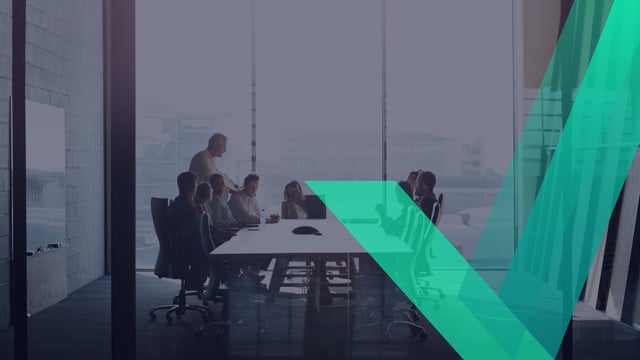 A group of people seated around a long conference table in a glass-walled meeting room, with one person standing and addressing the group.
